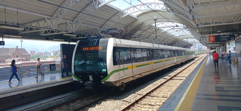 A white and green Medellín metro train pulling into Cisneros station under an arched roof